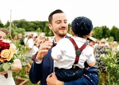 Père heureux portant son fils lors de la cérémonie de mariage, la mère à gauche brandit fièrement un bouquet de fleurs.